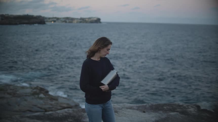 A woman stands on the shore holding a metal container in her arms and looks down at it