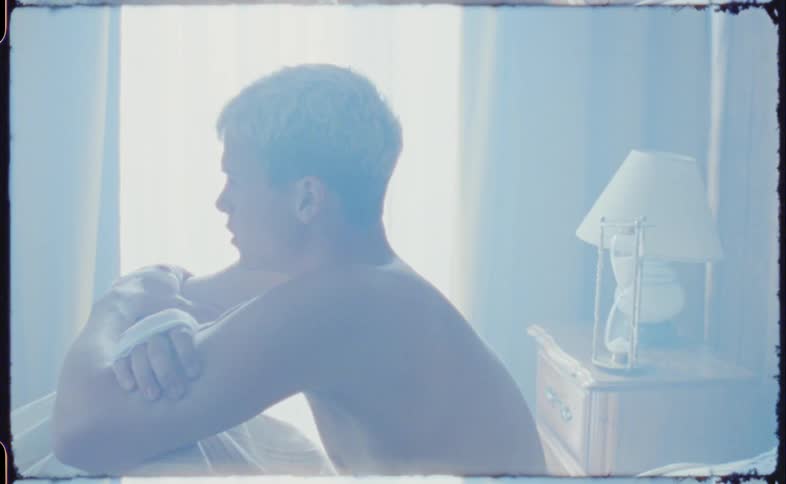 A shirtless young man sits and cries on his bed with his knees pulled to his chest