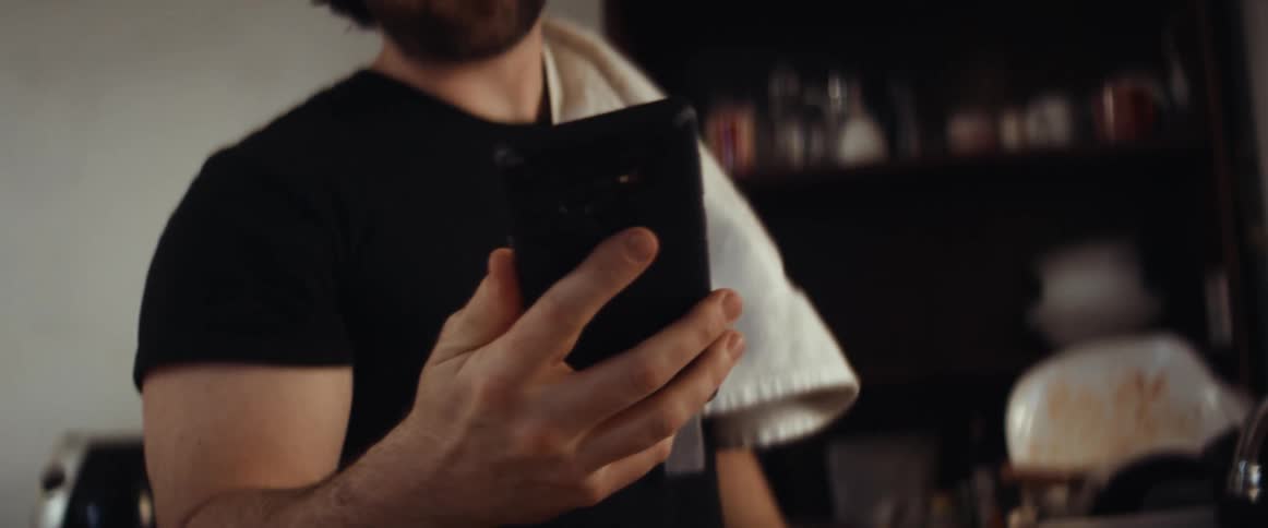 A man is standing in his kitchen and looking at a cell phone while he cooks