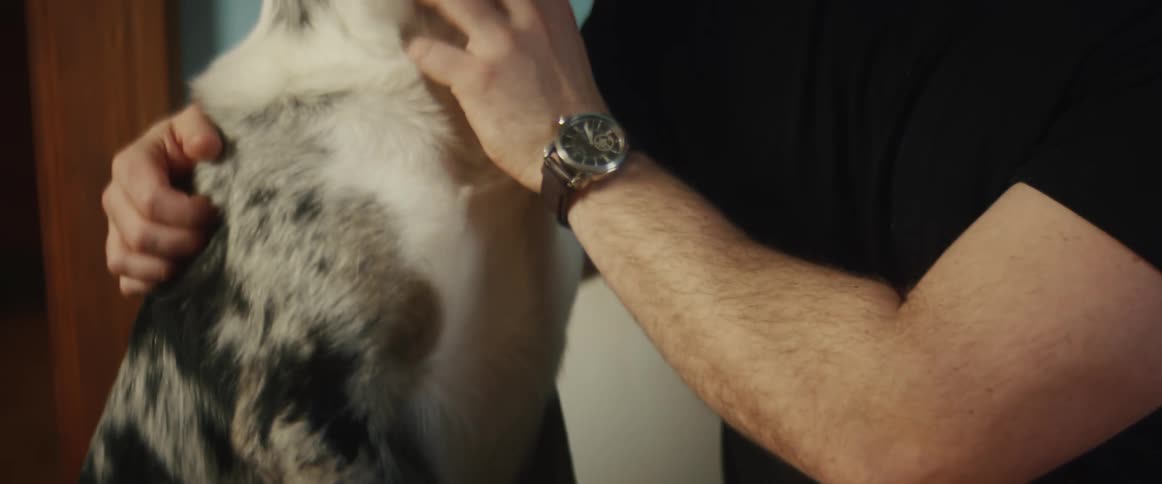 Man with a beard and a black t shirt pets a dog in front of turquoise wall