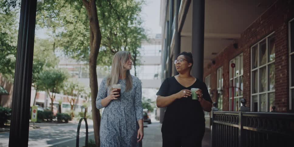 Two women walking holding cups with trees buildings the sky and clouds in the background