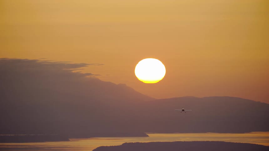 A single engine acrobatic airplane flies near mountains across an orange sky with a bright sunset on the horizon