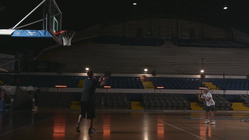 A man in a gym tossing a basketball to a woman as she shoots the ball at the hoop