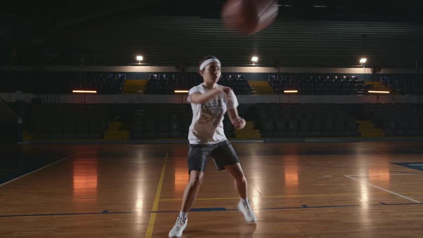 A girl with a headband in a gym practicing dribbling and passing a basketball with seats in the background