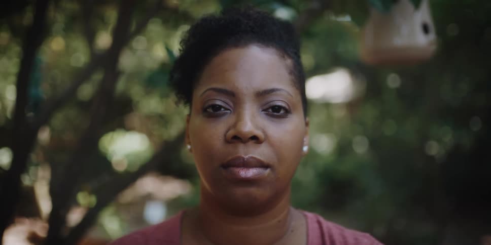 Woman with earrings and short hair faces the camera with eye contact in front of the trees