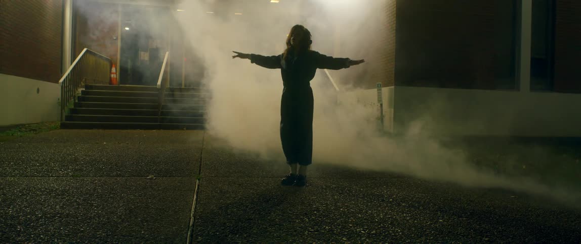 A woman dances hip hop style outside a building as a light flashes and thick smoke surrounds her