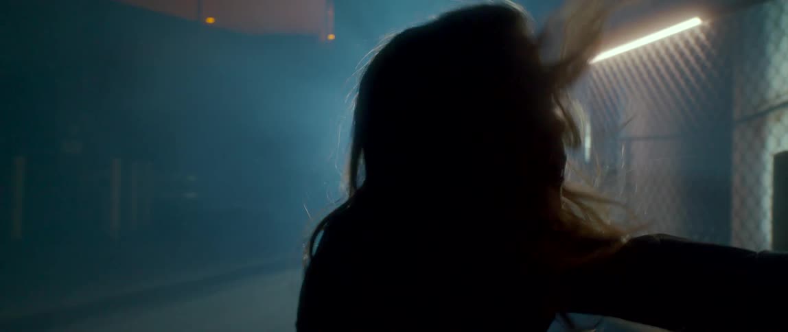 A woman dances outside at night in front of a fence with a beam of light shining through smoke in the distance