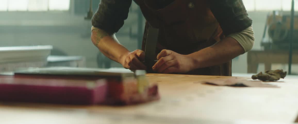 A man leans over a table as he uses a thin piece of sandpaper on a blade