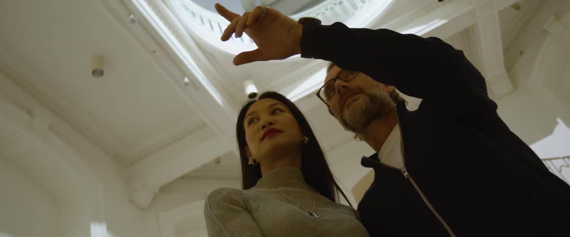 A man with glasses hugging and talking to a woman in a room with a staircase in the background