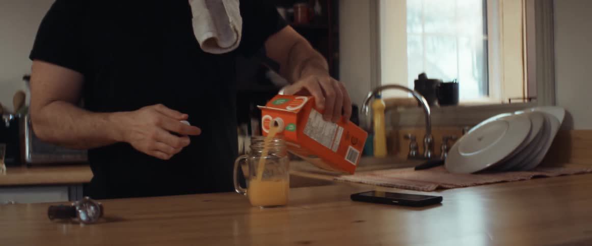 A man in a kitchen pouring orange juice into a glass with a doorway in the background