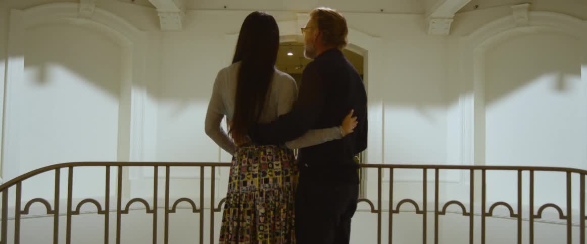 A man and woman come up two opposite stairs in a building and hug when they meet in the middle of the landing