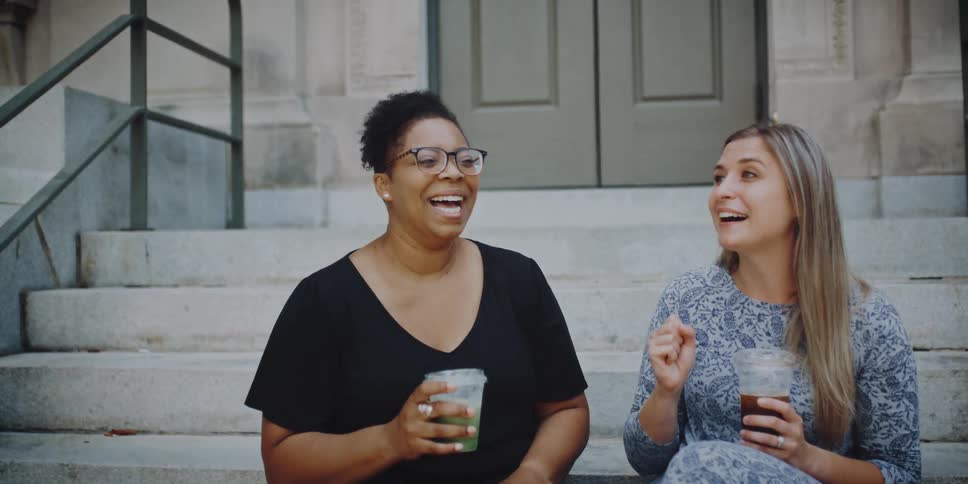 Two women sit on a set of stairs outside a building and talk while having non alcohol drinks