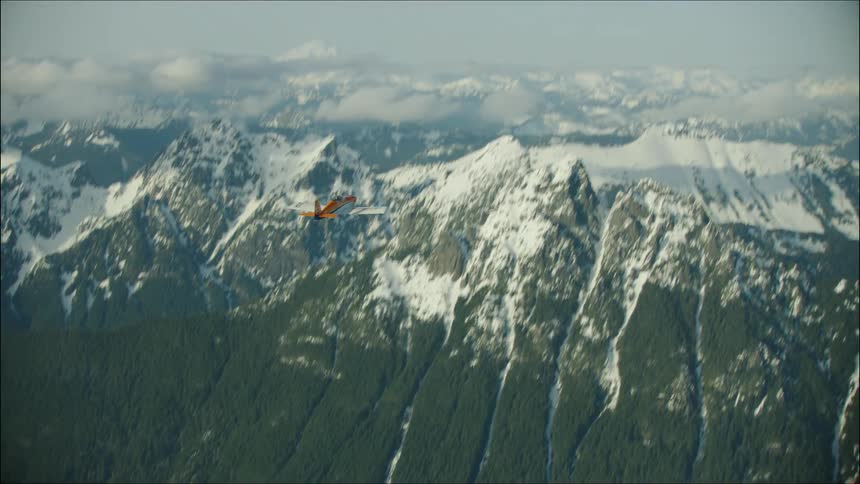 The single engine airplane quickly passes the camera revealing snow covered mountains before climbing rapidly into the blue afternoon sky