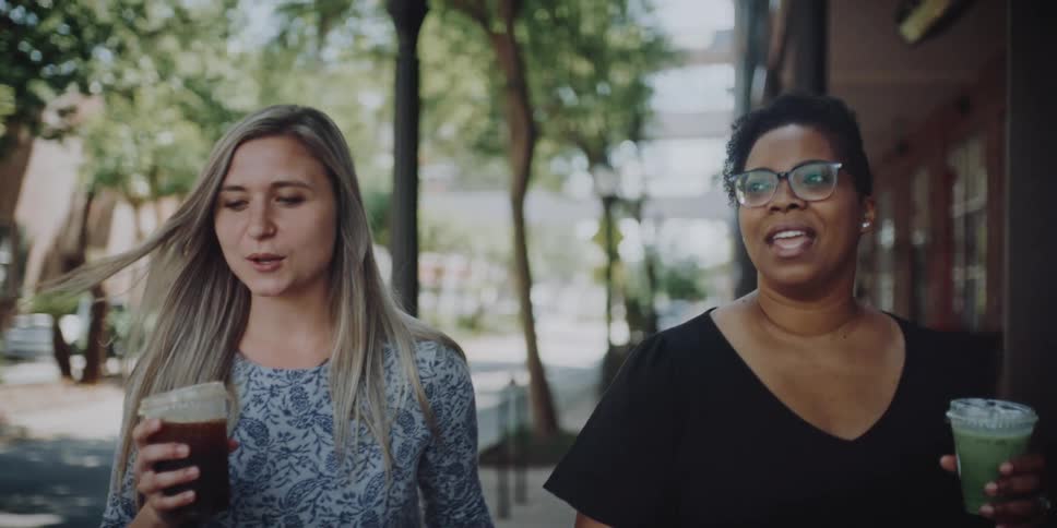 Blonde woman wearing a sweatshirt and black woman with glasses and a black t shirt holding plastic cups walk down the street