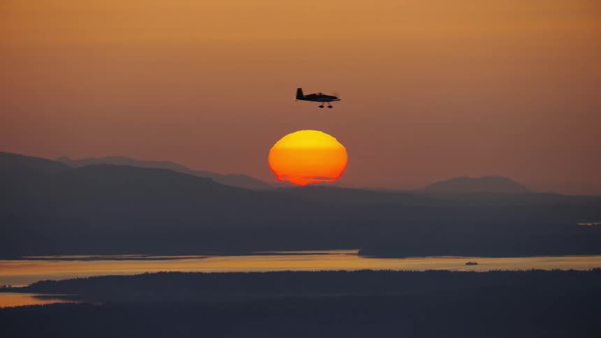 Epic end of day shot of an airplane flying over mountains across an orange sky with a bright sunset on the horizon