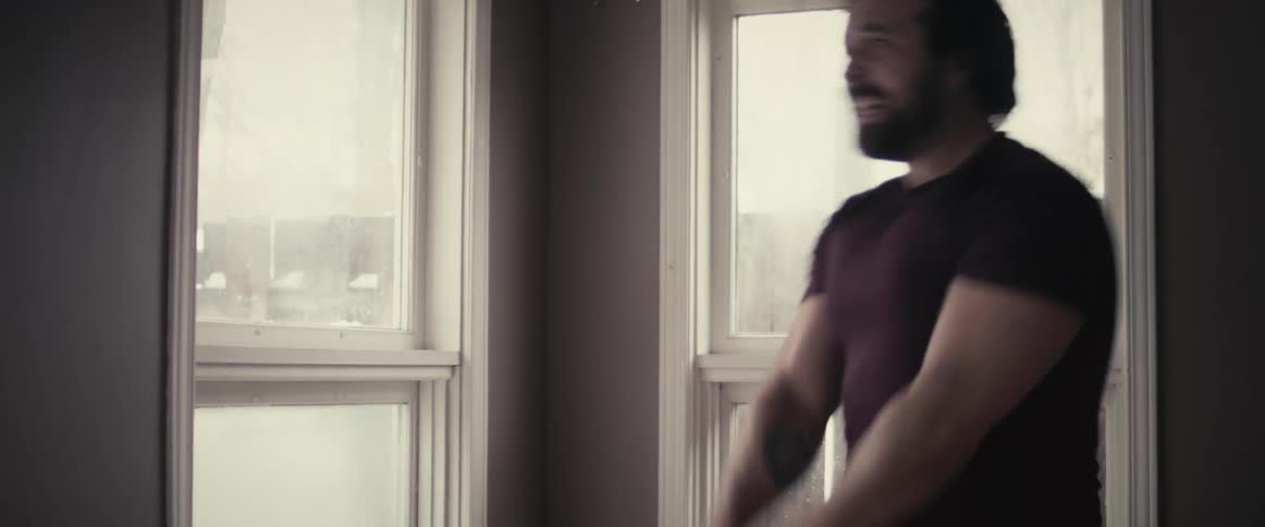 Muscular man with a t shirt and a beard lifts weights next to windows in the room