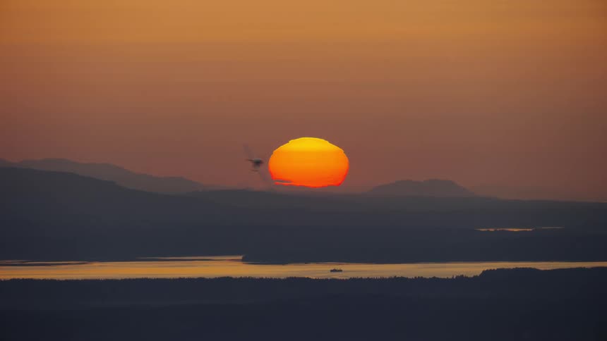 Airplane flies over rapidly oer the coastline across an orange sky with a bright sunset on the horizon and makes a sharp turn into frame