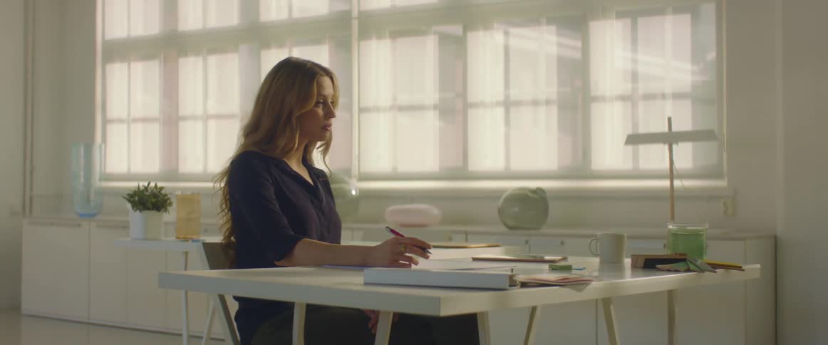A woman sitting on a chair at a desk with a wall and windows in the background