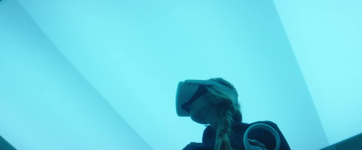 A woman wearing a virtual reality headset stands and waves a controller in front of herself