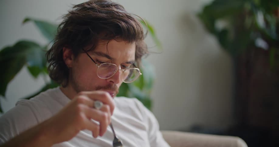 A man sits on the couch eating a bowl of food and looking at a laptop on his lap