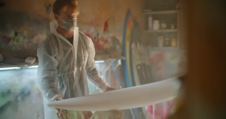 A man wearing protective dust gear places a unfinished surfboard on a saw horse