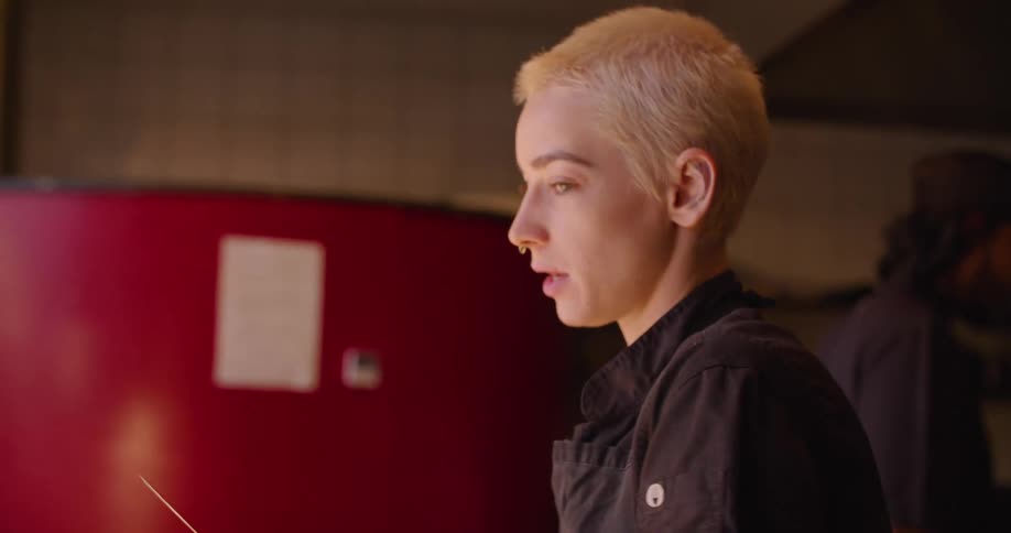 Blonde woman with short hair a nose ring a black shirt and apron cuts vegetable on a green cutting board in the restaurant kitchen
