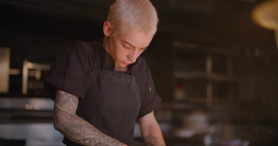 Blonde woman with short hair a black apron and tattoo cuts meat on the cutting board in the restaurant kitchen