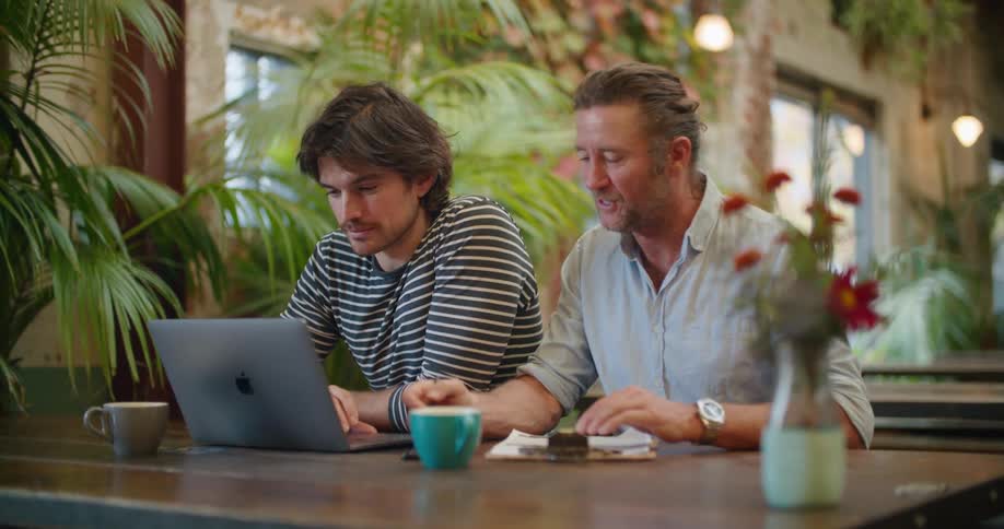 Two men sit in the restaurant looking at a laptop and talking