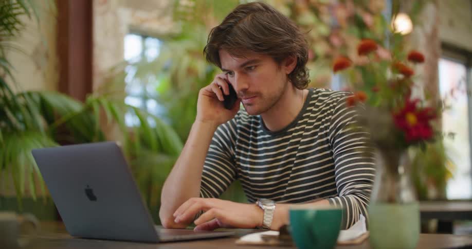 A man sitting at a desk talking on a smartphone and using a laptop with plants in the background