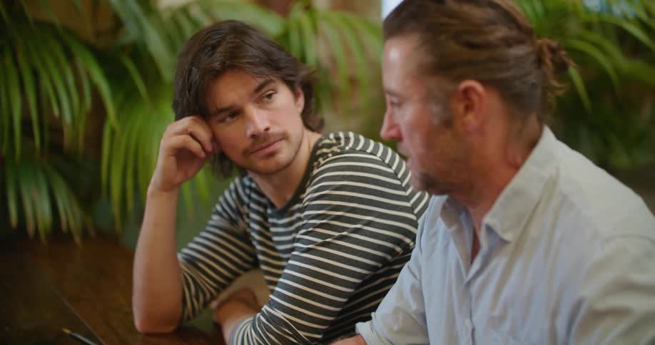 Two men sitting at a desk with a laptop with plants and a wall in the background