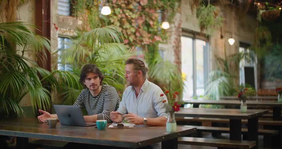 Two men sit at table near laptop in empty restaurant having a discussion