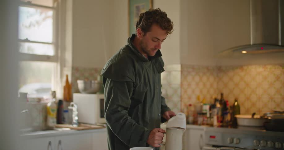A man in a kitchen placing a container on a table with a wall and window in the background