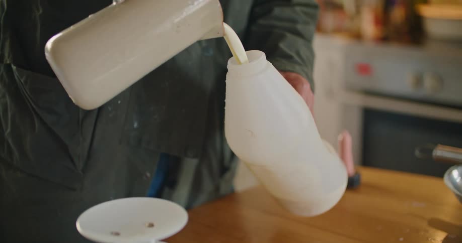 A person in a kitchen pouring milk into a bottle over a table