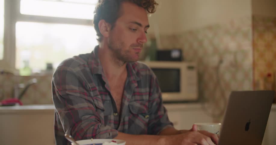 A man sits at a kitchen table typing on a laptop with a bowl beside him