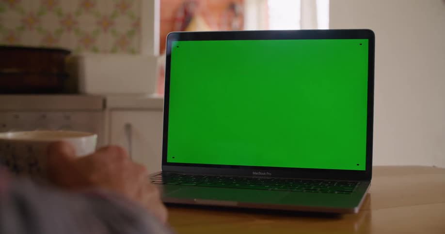 A man sitting at a kitchen table grasps a mug in his right hand in front of a macbook pro displaying a green screen while natural light floods in from a room in the background