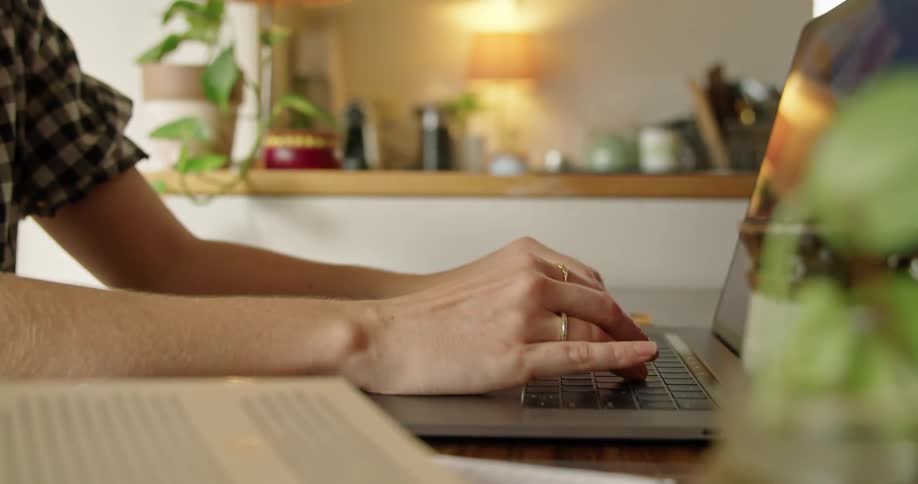 Woman with a plaid blouse and rings types on laptop keyboard in the kitchen at home