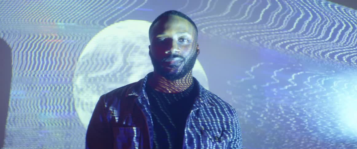 A man with a jacket standing in a room with a screen behind