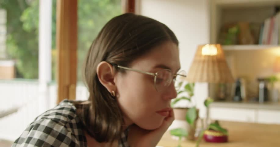 A woman with glasses sitting at a table using a laptop with a plant and a lamp in the background