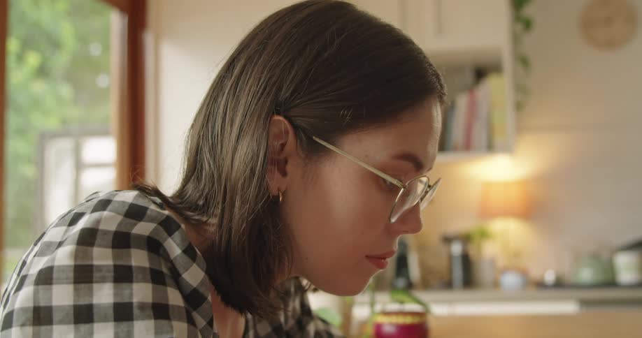 Woman with glasses and a plaid shirt sitting at table uses a laptop in the kitchen