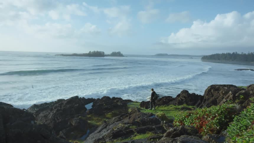 A man with a backpack holding a surfboard with the ocean sky and clouds in the background