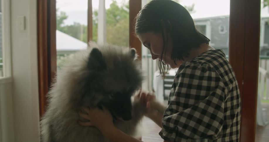 A woman kneels on the floor near the glass door of her home and plays with a large fluffy dog