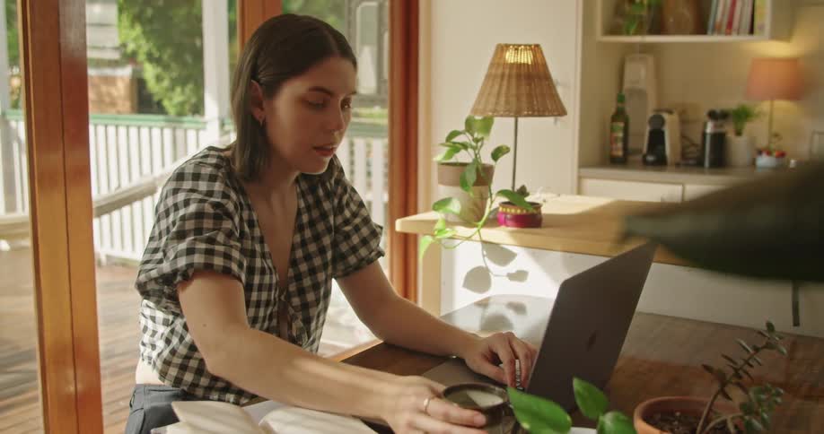 A woman sitting at a table next to a sliding door drinks from a coffee cup while working on a laptop computer during the day
