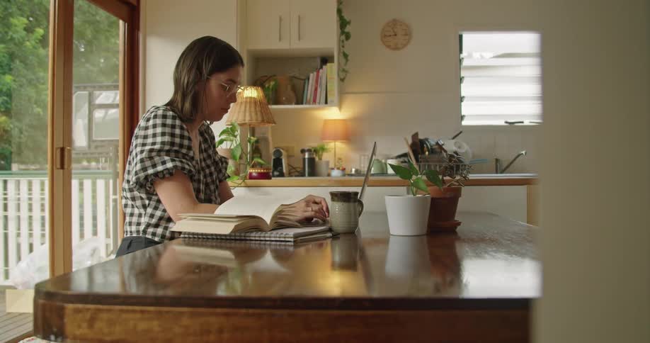 A woman sitting at a table at home next to a patio window typing on a laptop computer during the day