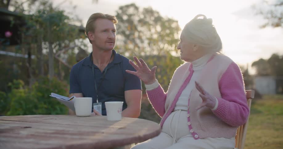 Man with a dark blue polo shirt and elderly woman wearing a pink sweater and a white vest sit at the table and talk to each other in the garden