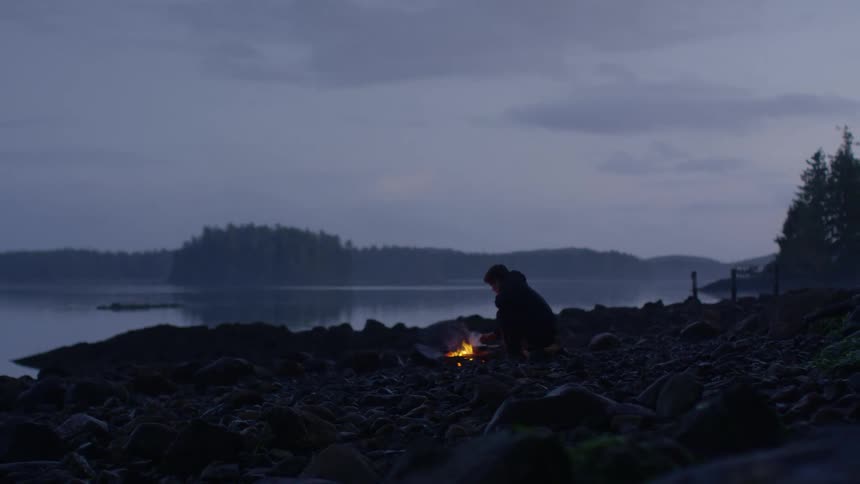 Male wearing dark colored clothes is sitting next to a campfire along the shore