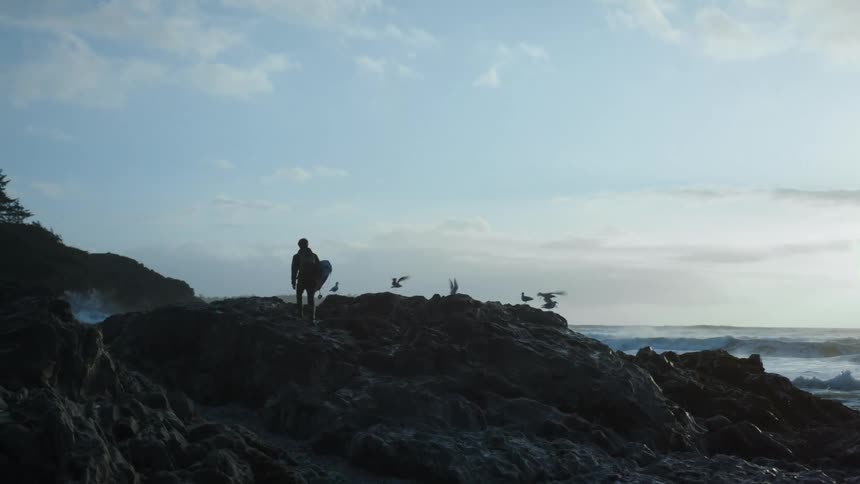 Silhouette of a man carrying a surfboardon the rocks next to the ocean at sunset