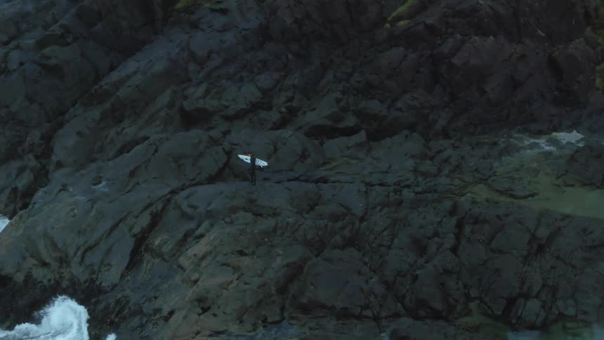 Male carrying a white surfboard walks across rocky terrain as waves splash against the rocks