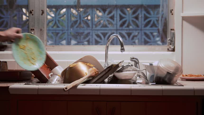 A person putting a dirty dish in a kitchen sink with a wall and window in the background