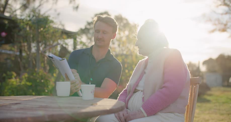 A man and elderly woman smiling while reading a book and drinking coffee outside on a sunny day