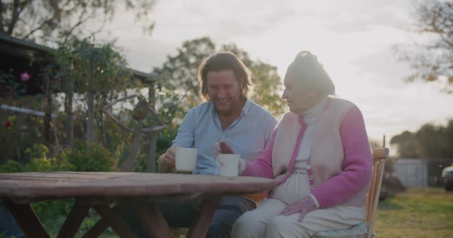 A man and older woman sit at a backyard table talking and drinking from small mugs